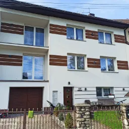 Dwukondygnacyjny dom jednorodzinny z jasną elewacją, brązowymi akcentami w postaci pasów i stolarki okiennej, ogrodzony metalowym płotem z kamiennymi słupkami.