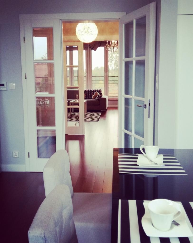 Białe, przeszklone drzwi dwuskrzydłowe otwarte do salonu z drewnianą podłogą i żyrandolem, widok z jadalni z nakrytymi stolikami.