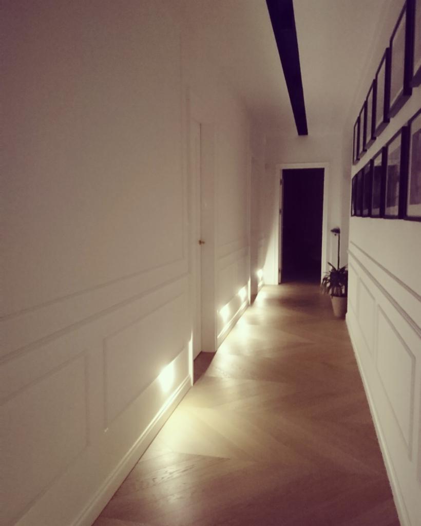 Korytarz z drewnianą podłogą ułożoną we wzór jodełki, białe ściany z panelami dekoracyjnymi, wpadające promienie słoneczne tworzą refleksy na podłodze.