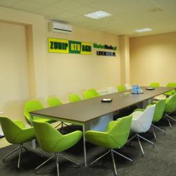 ZUHIP Sp. z o.o. - Jasna sala konferencyjna z długim stołem, zielonymi i białymi krzesłami obrotowymi, klimatyzacją i logotypem firmy na ścianie.