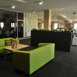 ZUHIP Sp. z o.o. - Nowoczesne biuro z zielonymi sofami i stolikiem kawowym w strefie relaksu, widok na przeszkloną salę konferencyjną i stanowiska pracy.