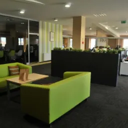 Nowoczesne biuro z zielonymi sofami i stolikiem kawowym w strefie relaksu, widok na przeszkloną salę konferencyjną i stanowiska pracy.