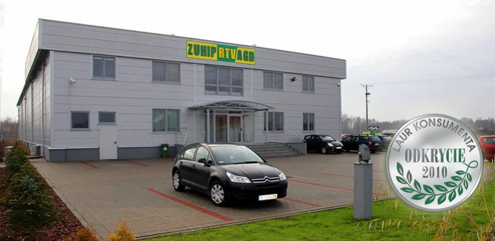 Szary budynek firmy ZUNIP RTVAGD z zielonym logo na fasadzie, czarny samochód zaparkowany przed wejściem, na pierwszym planie naklejka 'Laur Konsumenta - Odkrycie 2010'.