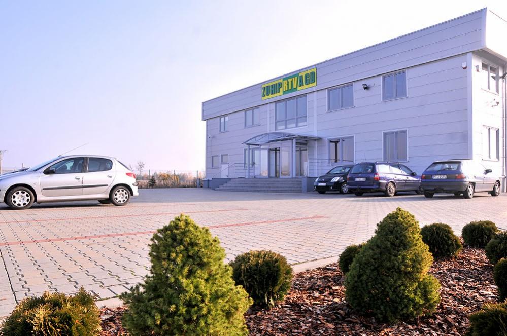 Budynek firmy ZUHIP RTV/AGD z zaparkowanymi samochodami i ozdobnymi krzewami na pierwszym planie.