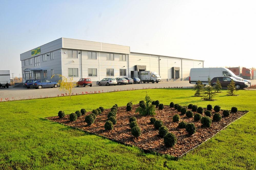 Widok z zewnątrz na nowoczesny budynek magazynowo-biurowy z parkingiem i zadbanym trawnikiem z ozdobnymi nasadzeniami.