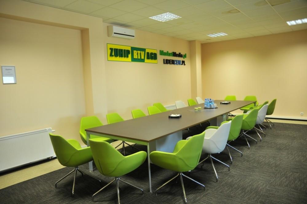 Jasna sala konferencyjna z długim stołem, zielonymi i białymi krzesłami obrotowymi, klimatyzacją i logotypem firmy na ścianie.