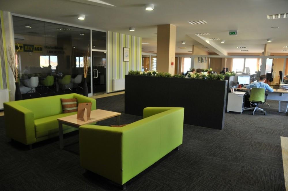 Nowoczesne biuro z zielonymi sofami i stolikiem kawowym w strefie relaksu, widok na przeszkloną salę konferencyjną i stanowiska pracy.