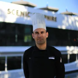 Portret kucharza w stroju firmowym na tle luksusowego statku wycieczkowego z napisem 'Scenic Sapphire'.