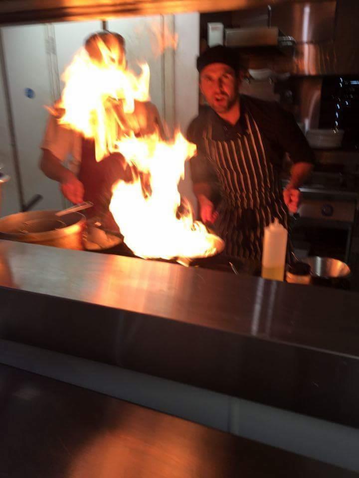 Kucharz w czapce i pasiastej bluzie flambirowuje potrawę na metalowej patelni, z tyłu drugi kucharz w tle kuchni.
