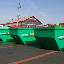 Rząd zielonych, pustych kontenerów na gruz firmy MAXICAR, ustawionych na parkingu z kostki brukowej, z budynkiem firmy w tle.