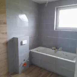 Szara łazienka w trakcie wykańczania, widoczna wanna obudowana płytkami, zabudowa stelaża WC i fragment podłogi z płytek imitujących drewno, surowy stan instalacji elektrycznej.