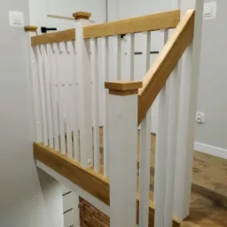 Nowoczesna balustrada schodowa z jasnego drewna i białych pionowych listew, widok z góry na dół, z widoczną ścianą z cegły klinkierowej na dole.