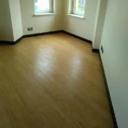 Jasne panele podłogowe ułożone diagonalnie w rogu pokoju z dwoma oknami i ciemnymi listwami przypodłogowymi.