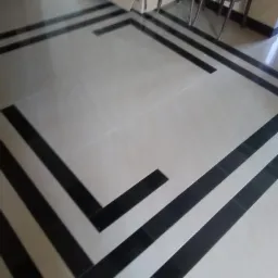 Ułożona podłoga z jasnych płytek z czarnymi dekoracyjnymi pasami, widok z góry, w tle karton i krzesła.