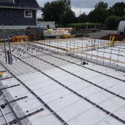 Przygotowanie do wylewki fundamentu: ułożona izolacja termiczna, siatka zbrojeniowa i rury ogrzewania podłogowego na warstwie styropianu, widoczne elementy instalacji hydraulicznej i elektrycznej...