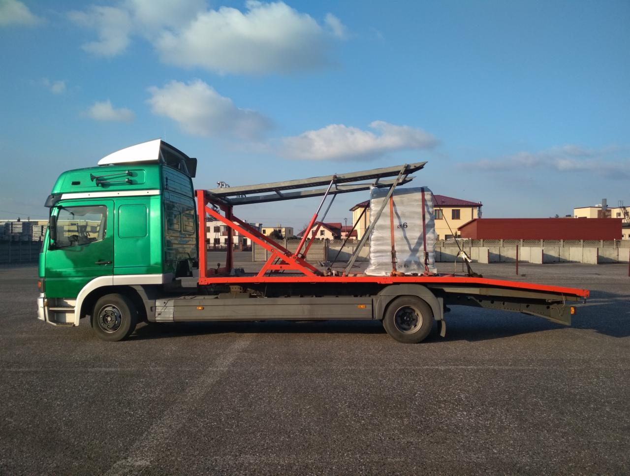 Zielona laweta z czerwonym stelażem przewozi zabezpieczony ładunek na otwartym parkingu w słoneczny dzień.