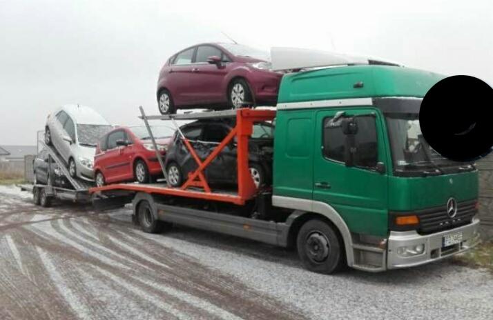 Zielona laweta Mercedes z trzema samochodami osobowymi różnych marek i kolorów na platformie, transportowana po szarej drodze w pochmurny dzień.
