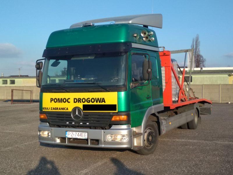 Zielono-srebrna laweta z napisem 'Pomoc Drogowa Kraj-Zagranica' na żółtym tle, zaparkowana na placu. Widoczne tablice rejestracyjne i elementy konstrukcyjne platformy transportowej.