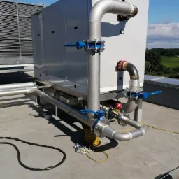 Instalacja przemysłowa na dachu budynku, widoczne rury, zawory z niebieskimi uchwytami, elementy spawane i klimatyzatory przemysłowe w tle.