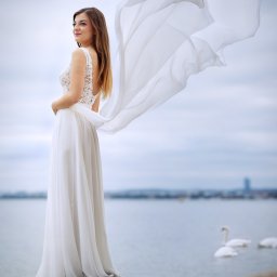 Nadolny Photography - Panna młoda w koronkowej sukni, stojąca na skale z powiewającym welonem na tle jeziora z łabędziami.