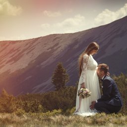 Nadolny Photography - Przyszły pan młody w granatowym garniturze całuje brzuszek panny młodej w koronkowej sukni na tle górskiego krajobrazu.