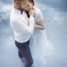 Nadolny Photography - Para młoda w objęciach na tle rozmytego, błękitnego tła; on w białej koszuli i bordowej muszce, ona w koronkowej sukni i złotych kolczykach.