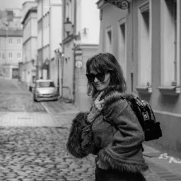 Czarno-białe zdjęcie kobiety w okularach przeciwsłonecznych, ubranej w sweter z futrzanymi mankietami i plecak, stojącej na brukowanej uliczce miejskiej.