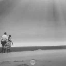 Czarno-białe zdjęcie pary idącej po piaszczystej wydmie w kierunku morza, pod promieniami słońca przebijającymi się przez chmury.