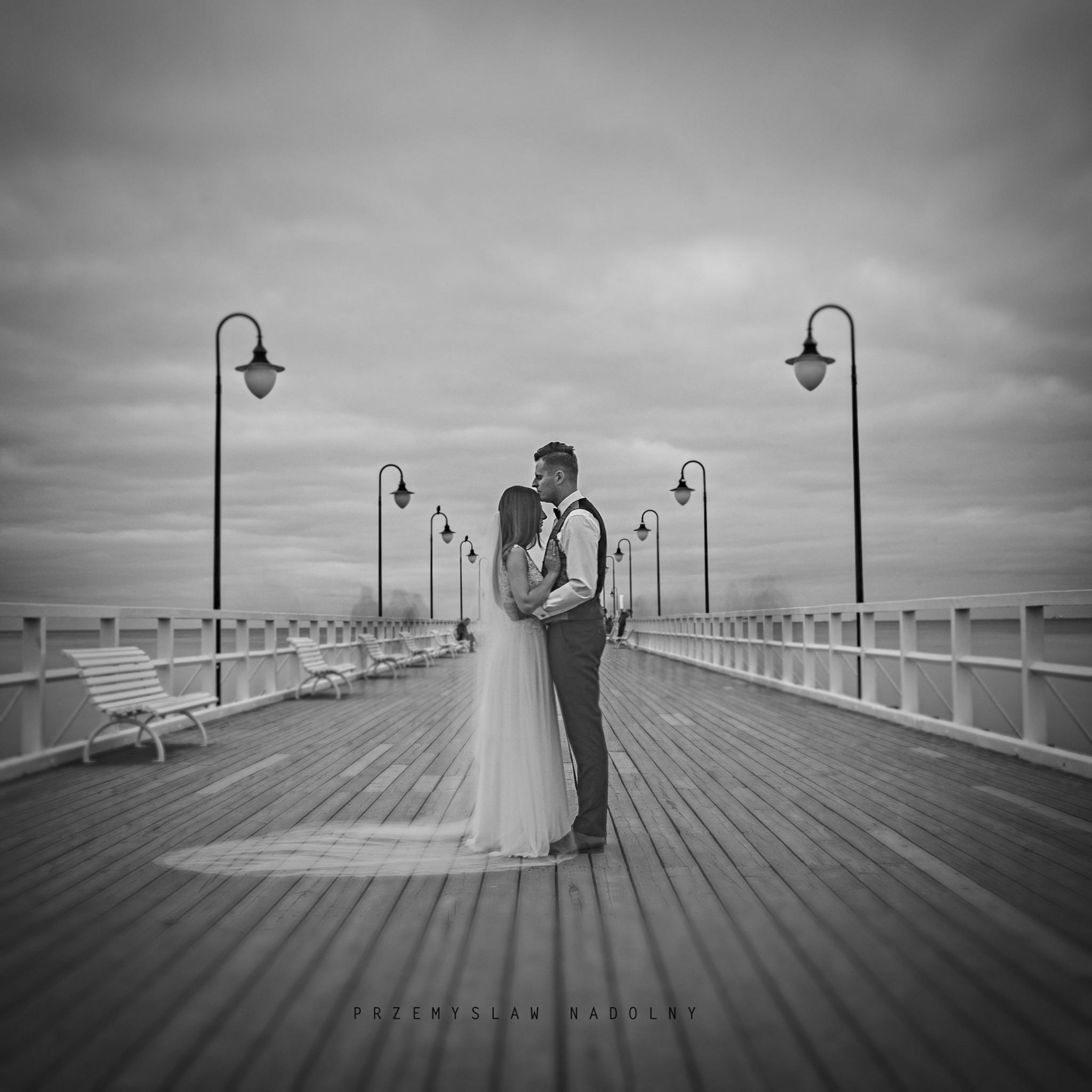 Czarno-białe zdjęcie pary młodej na molo z latarniami i ławkami, mężczyzna całuje kobietę w czoło, pochmurne niebo w tle.