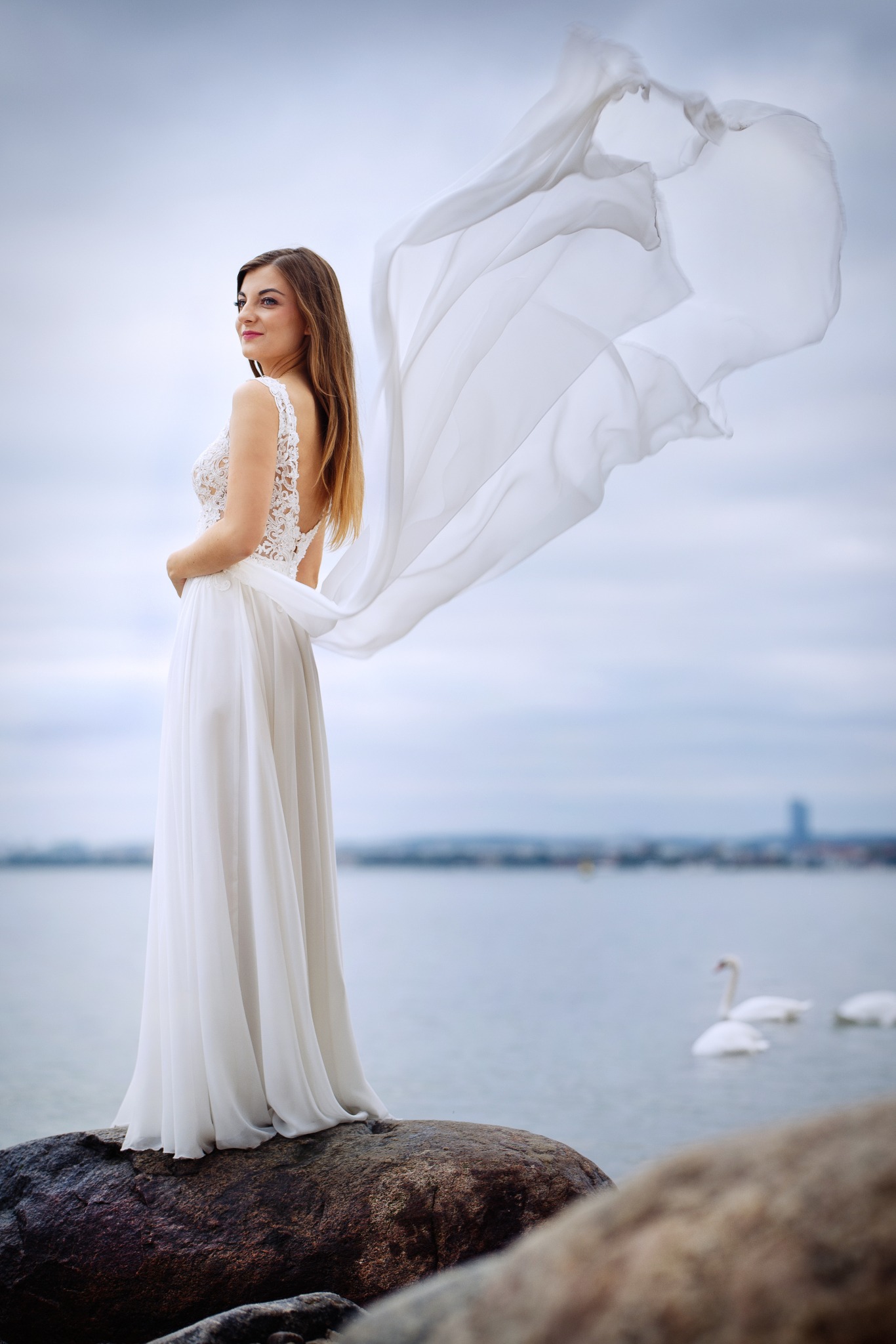 Panna młoda w koronkowej sukni, stojąca na skale z powiewającym welonem na tle jeziora z łabędziami.