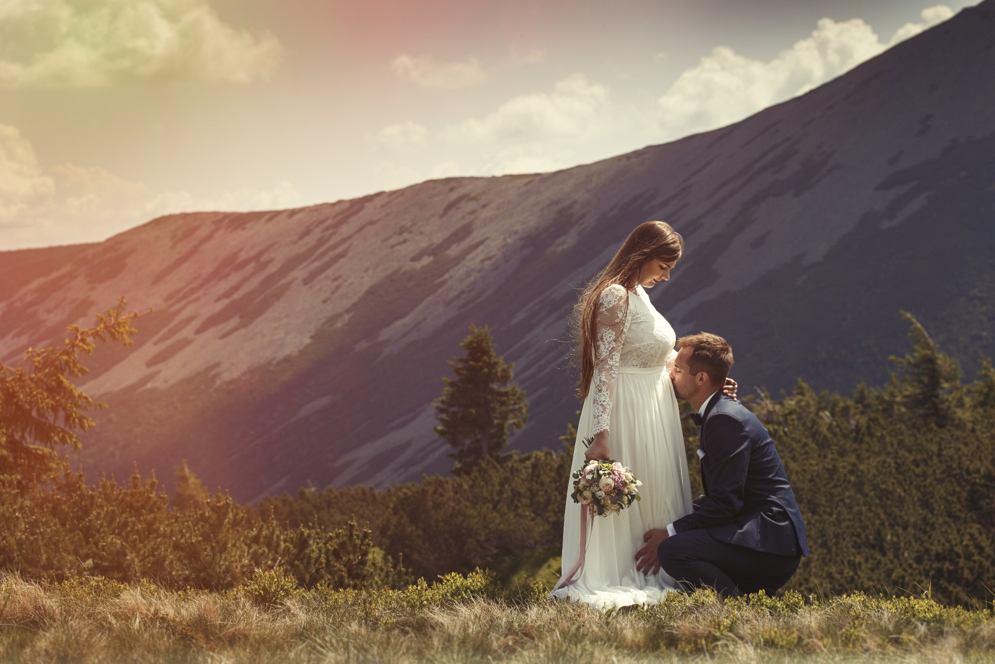 Przyszły pan młody w granatowym garniturze całuje brzuszek panny młodej w koronkowej sukni na tle górskiego krajobrazu.