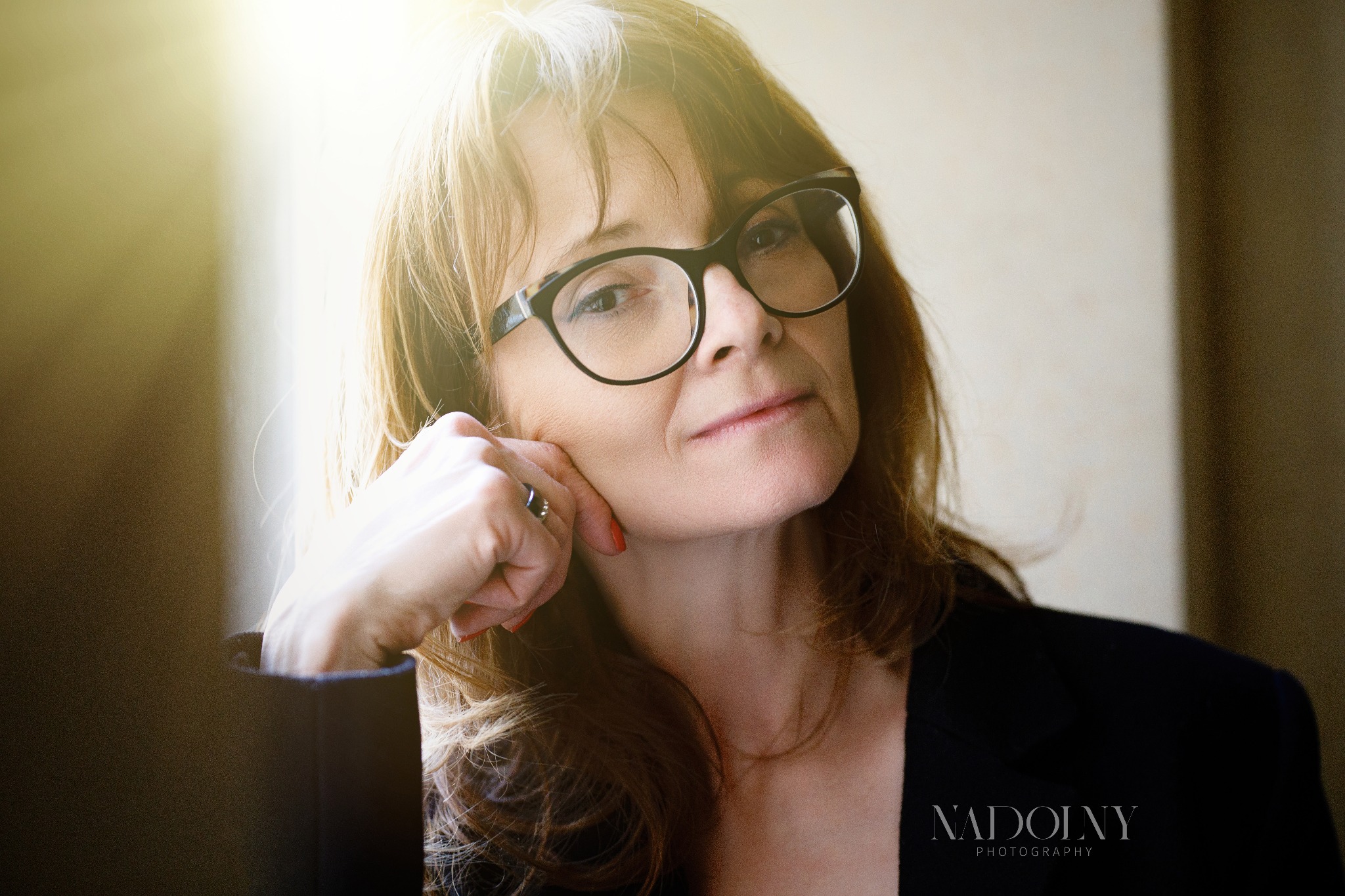 Portret kobiety w okularach, opierającej brodę na dłoni, z widocznym logo fotografa w prawym dolnym rogu.