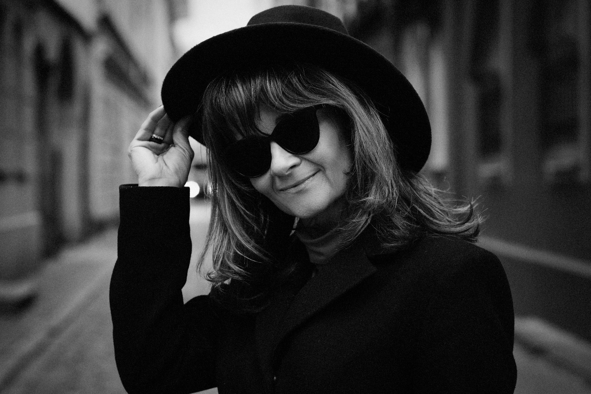 Portret kobiety w czarnym kapeluszu i okularach przeciwsłonecznych, uśmiechniętej, w miejskiej scenerii, czarno-biały.