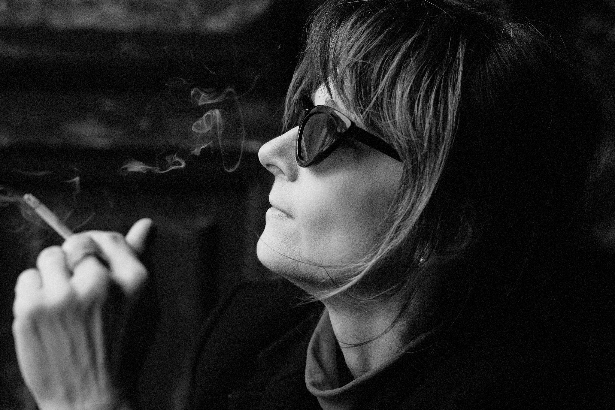 Czarno-białe zdjęcie profilu kobiety w okularach przeciwsłonecznych, palącej papierosa, z wyraźnym dymem unoszącym się w powietrzu.
