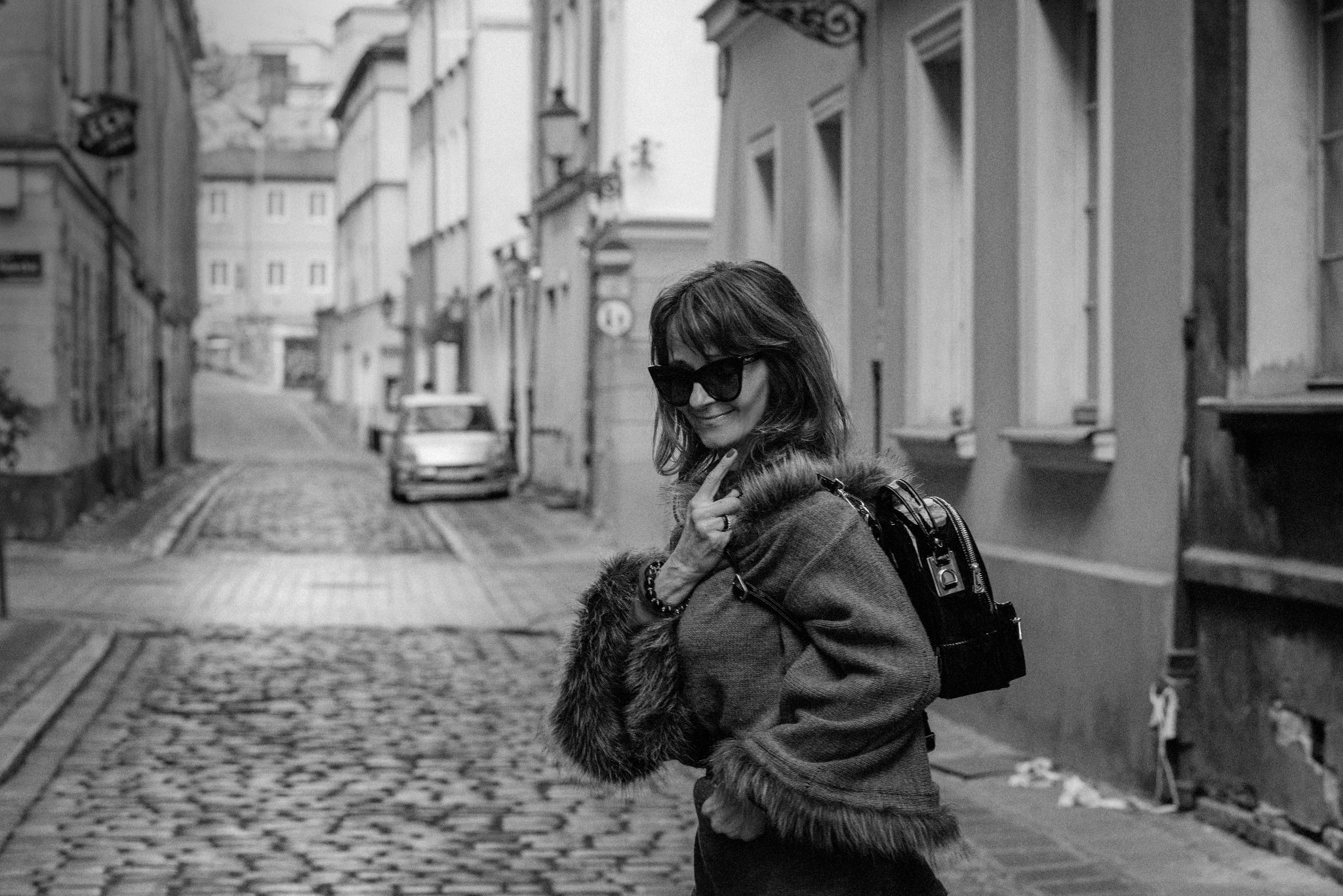 Czarno-białe zdjęcie kobiety w okularach przeciwsłonecznych, ubranej w sweter z futrzanymi mankietami i plecak, stojącej na brukowanej uliczce miejskiej.
