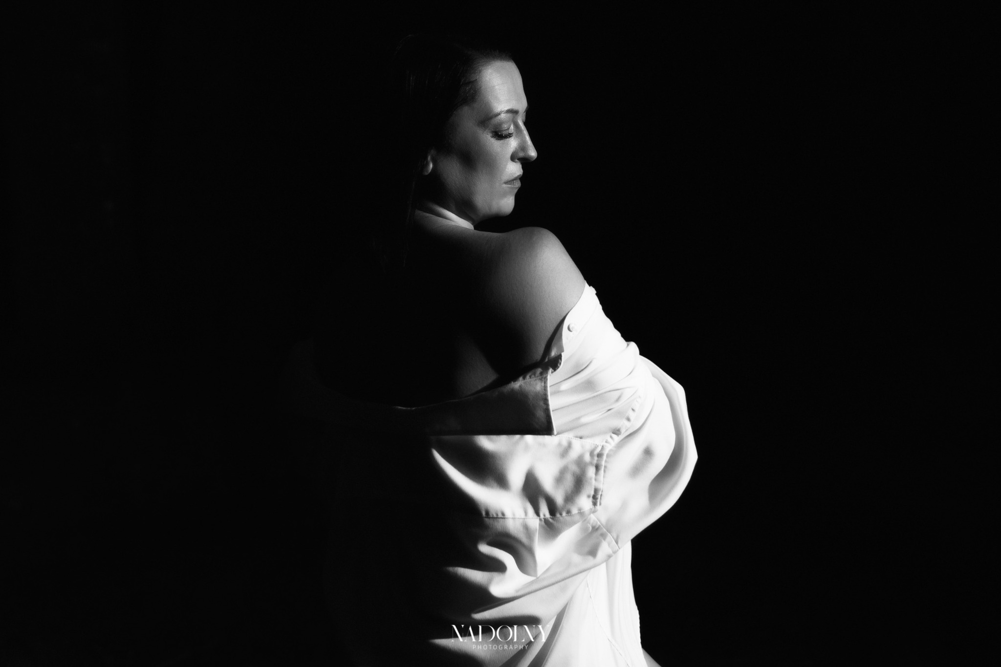 Czarno-białe zdjęcie kobiety w białej, rozpiętej koszuli, z zamkniętymi oczami i głową odwróconą w bok, na ciemnym tle.