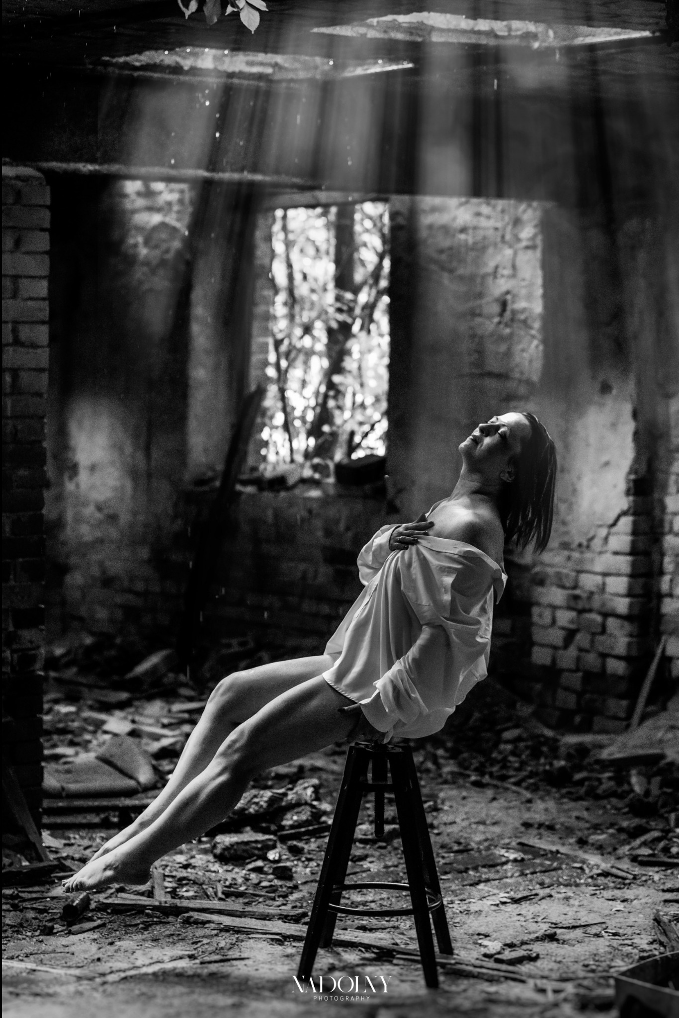 Kobieta w rozpiętej białej koszuli siedzi na stołku w opuszczonym, ceglanym pomieszczeniu, z promieniami światła wpadającymi przez okno, czarno-biała fotografia.