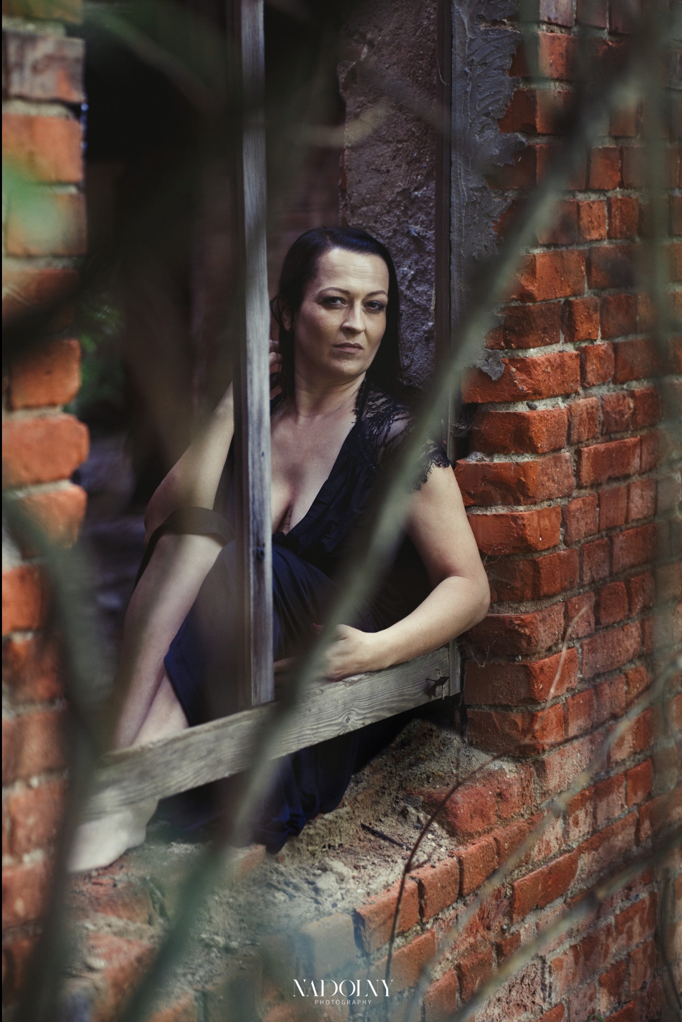 Kobieta w czarnej sukience z koronką siedzi w zrujnowanym oknie z cegły, spoglądając w obiektyw przez gałęzie na pierwszym planie.