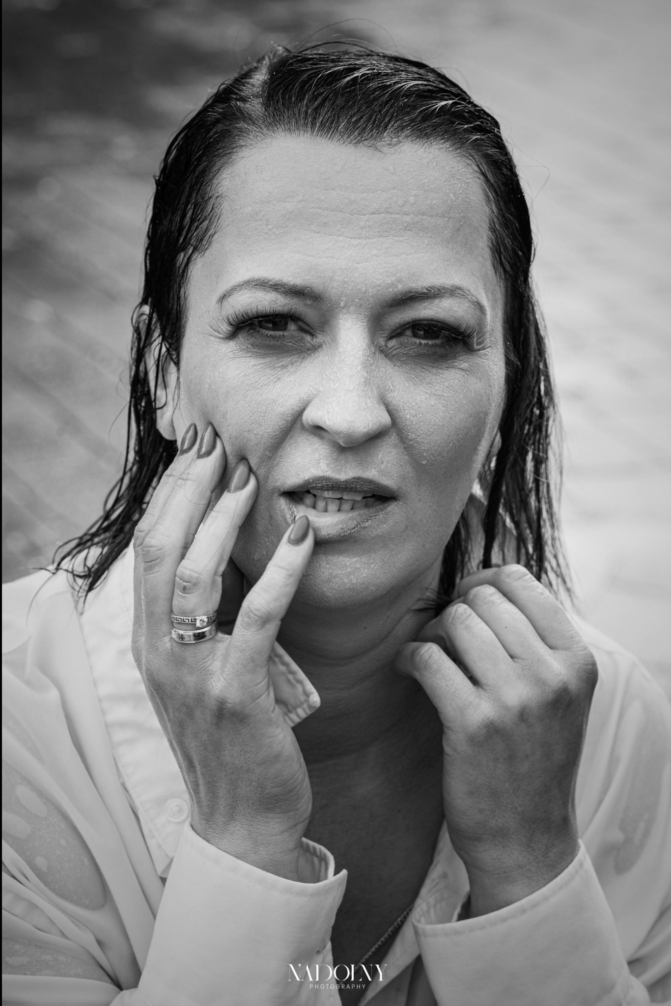 Portret kobiety z mokrymi włosami w rozpiętej białej koszuli, czarno-białe zdjęcie z bliska, dłoń z pierścionkiem na policzku, delikatny uśmiech.