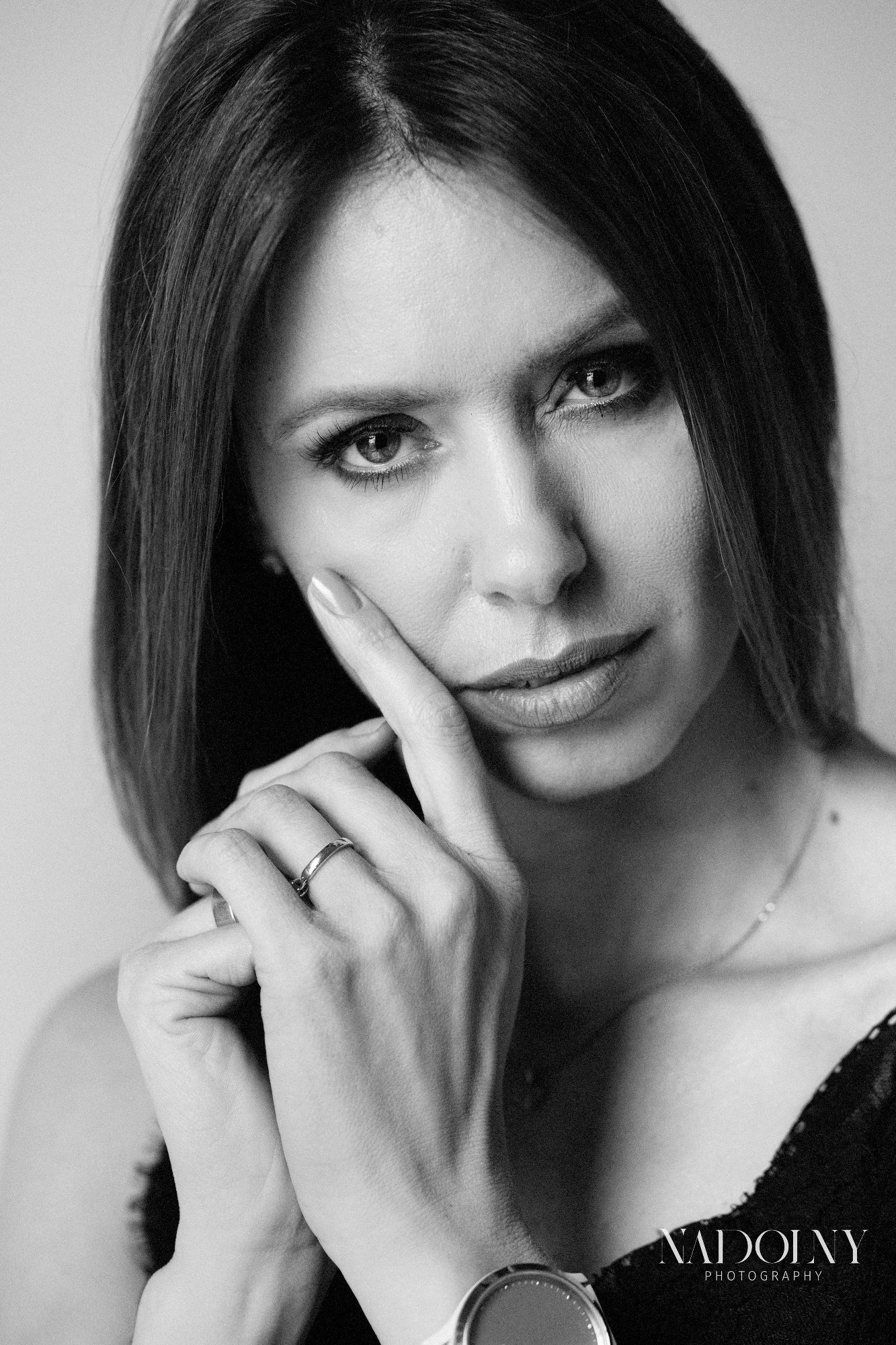 Portret kobiety w czerni i bieli, z pierścionkiem na palcu, opierającej brodę na dłoni, z delikatnym uśmiechem.