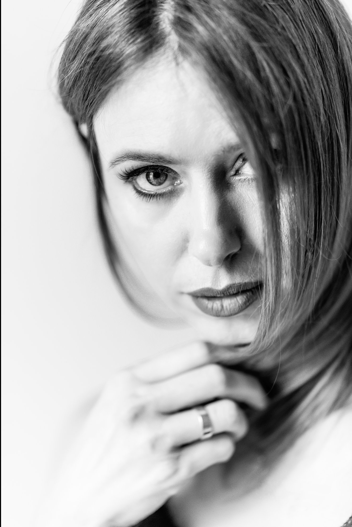 Portret kobiety w czerni i bieli, z jednym okiem wyraźnie widocznym, włosy opadają na twarz, dłoń z pierścionkiem pod brodą, intensywne spojrzenie.