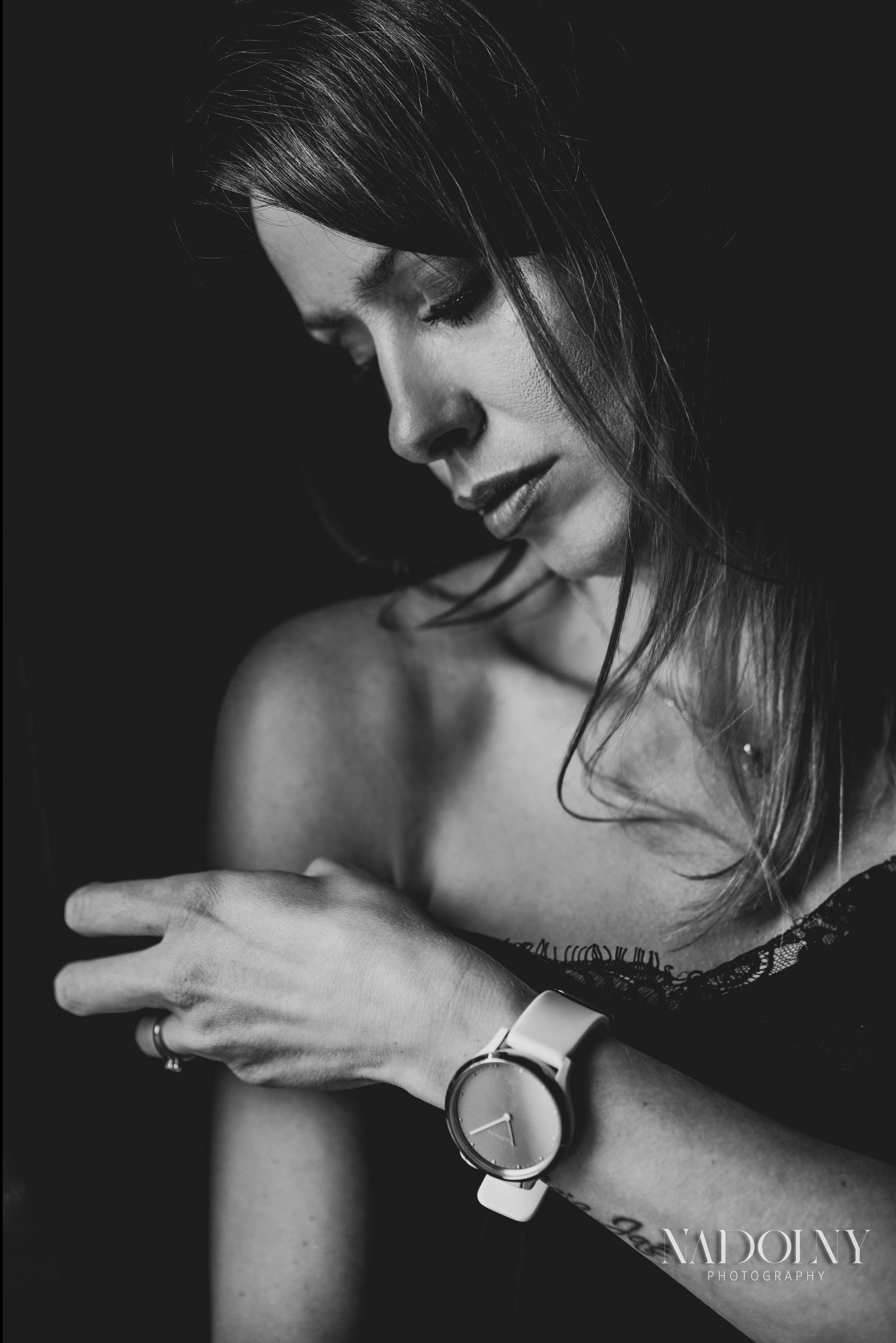 Portret kobiety w czerni i bieli, z delikatnym makijażem, zegarkiem na nadgarstku i tatuażem, spoglądającej w dół w miękkim oświetleniu.