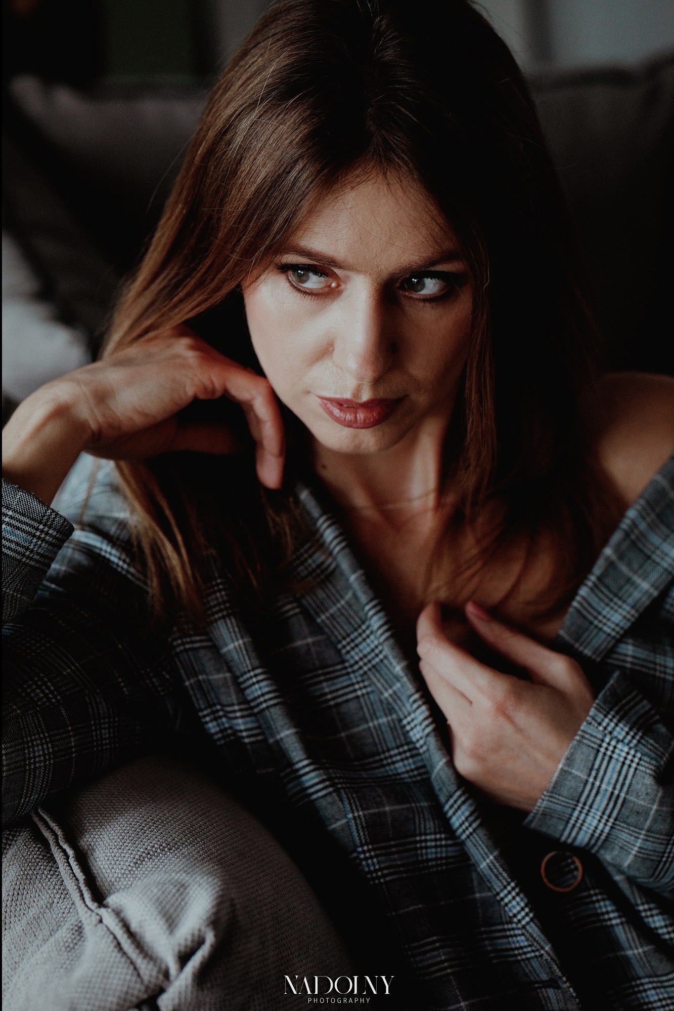 Portret kobiety w rozpiętej, kraciastej marynarce, zamyślona poza, delikatny makijaż, stonowane oświetlenie.