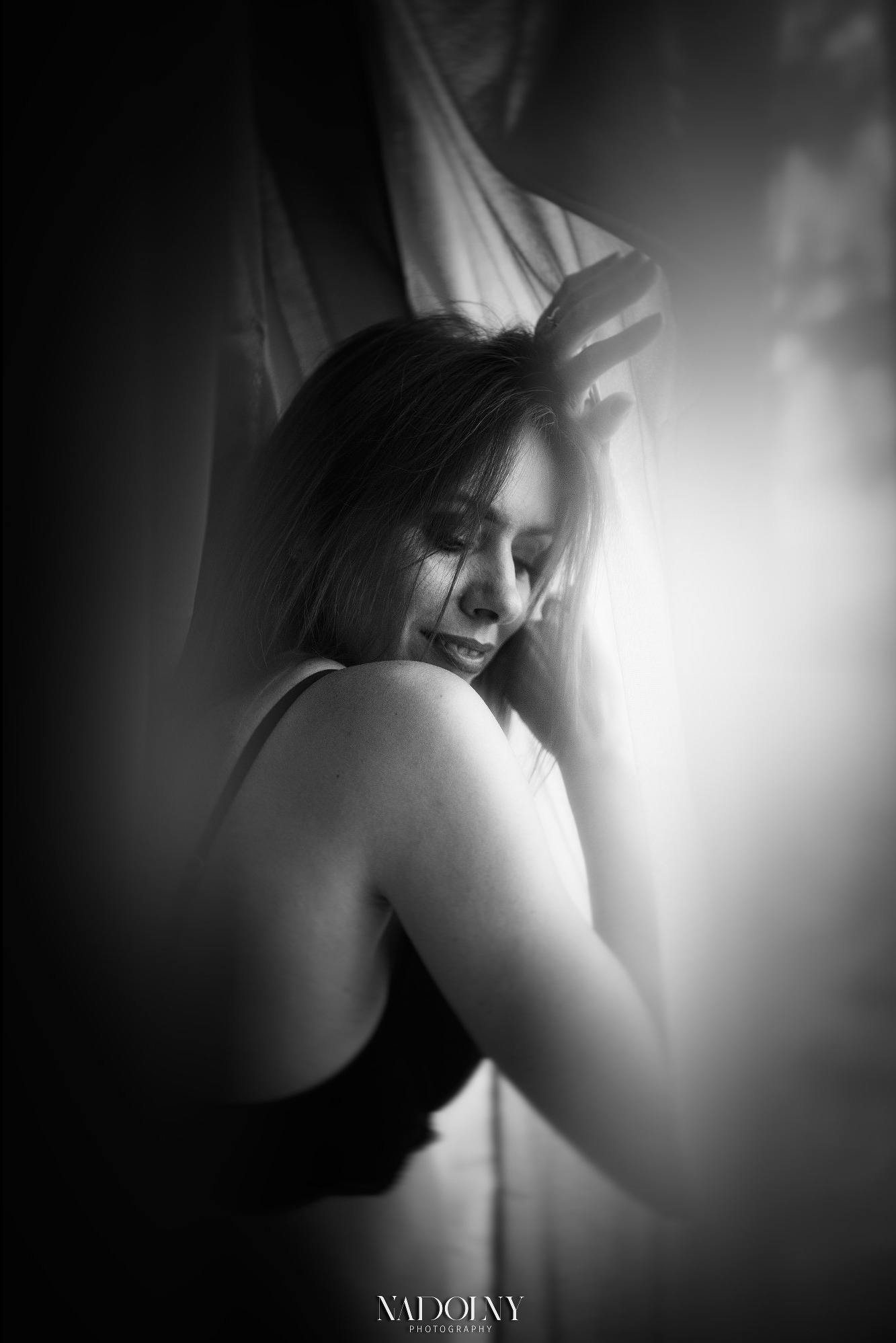 Portret kobiety w bieliźnie, z delikatnym uśmiechem, uchwycony w czerni i bieli, z subtelnym światłem padającym przez zasłony, podkreślającym jej naturalne piękno.