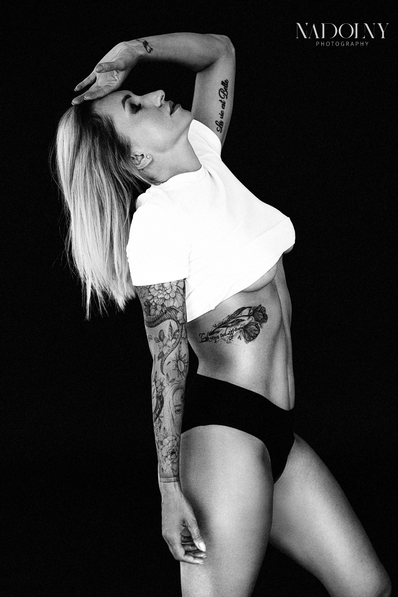Czarno-białe zdjęcie kobiety z tatuażami w białym topie i czarnych majtkach na czarnym tle, z ręką uniesioną do głowy, ukazujące profil i część ciała.