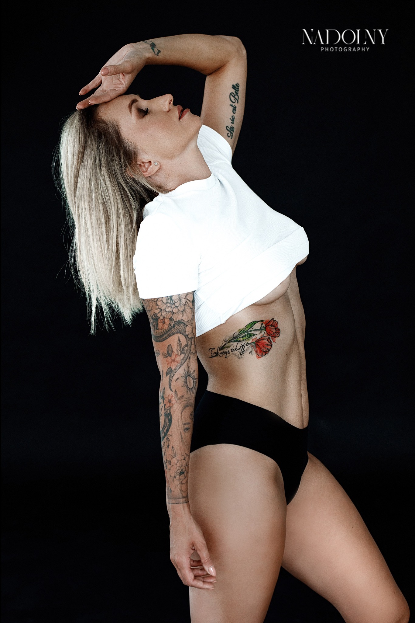Portret kobiety z blond włosami, w białym t-shircie i czarnych majtkach, z tatuażami na ramionach i boku, pozującej na czarnym tle