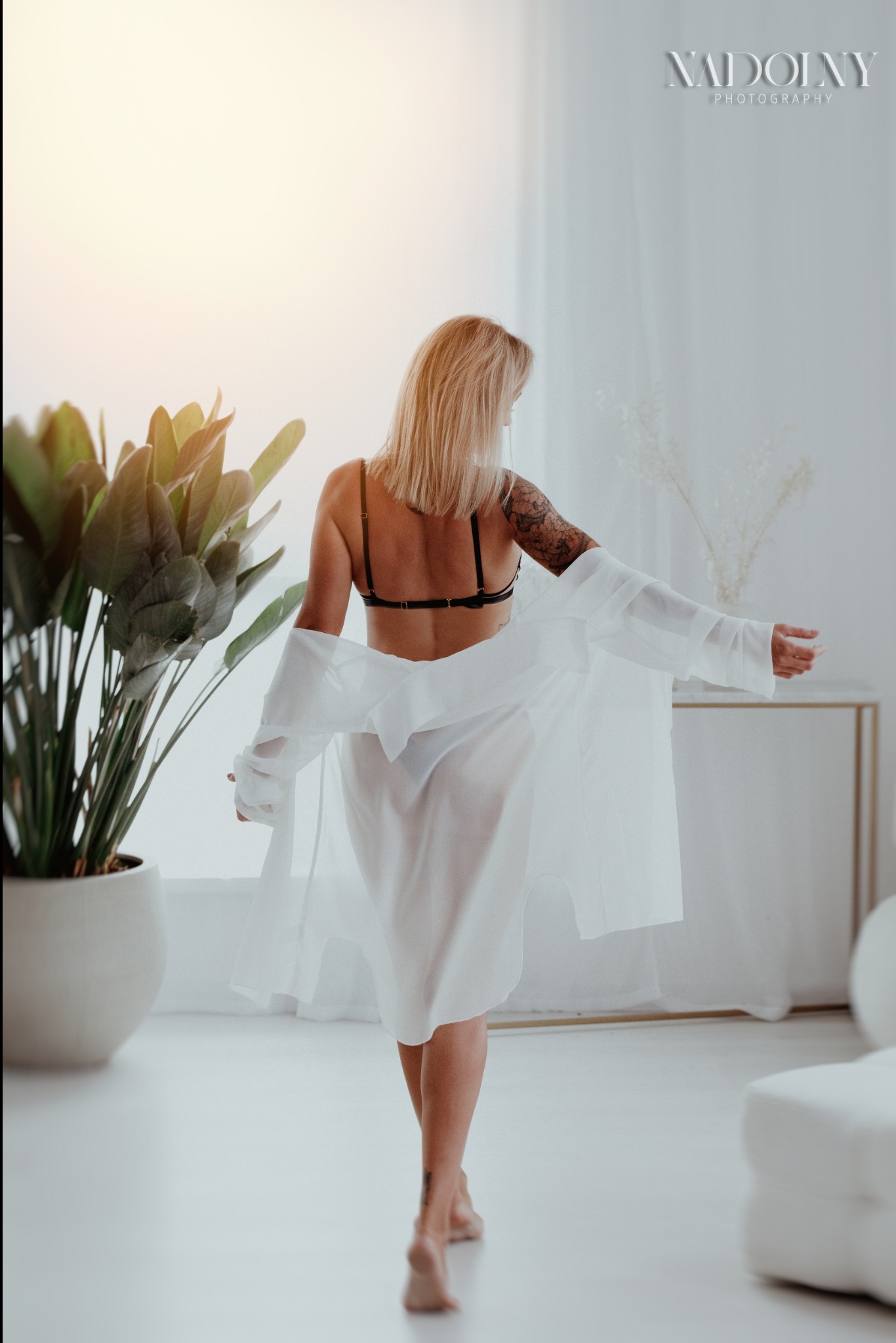 Kobieta odwrócona tyłem, w bieliźnie i rozpiętej białej koszuli, z tatuażem na ramieniu, idąca boso po jasnej podłodze w jasnym wnętrzu z rośliną doniczkową.