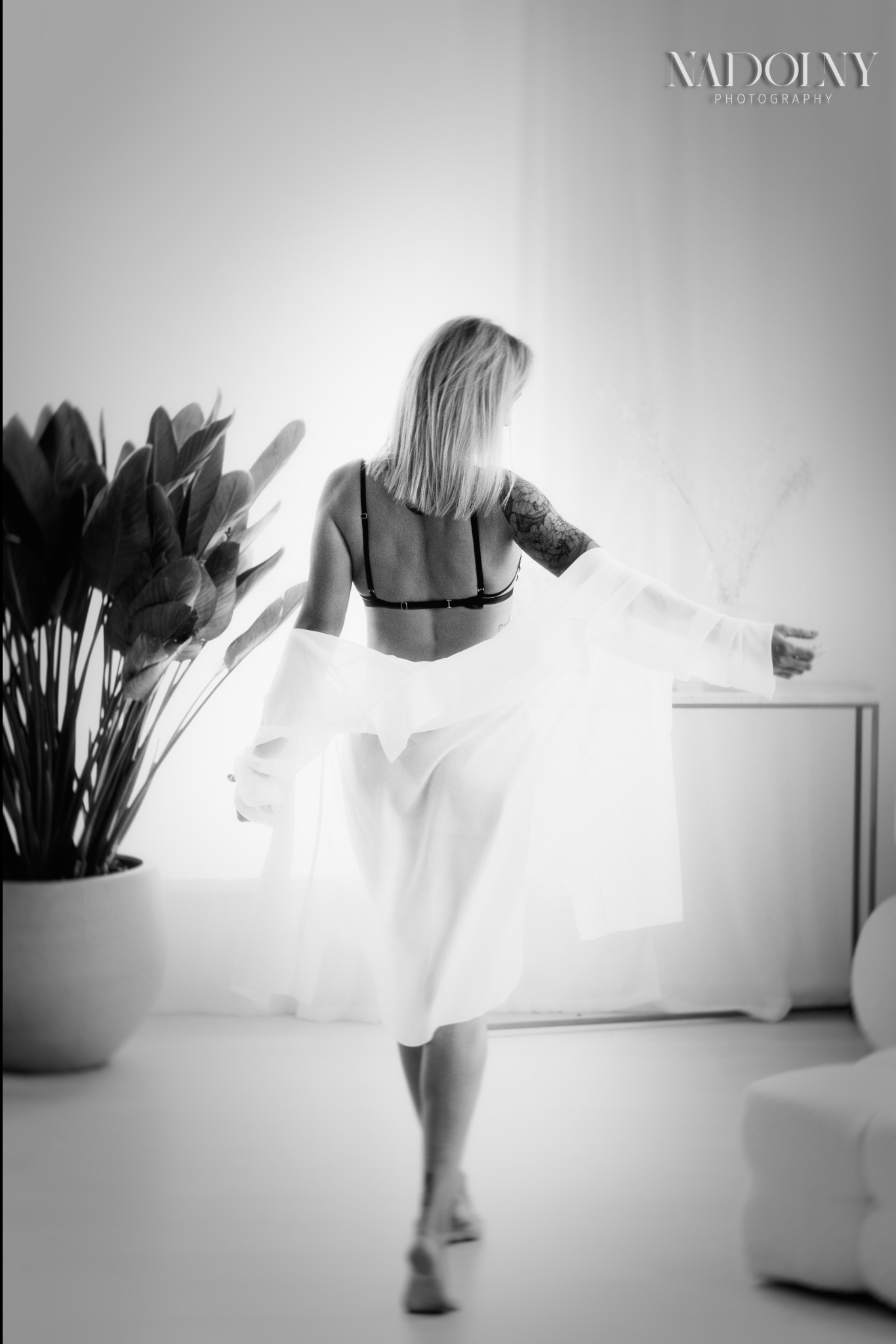 Czarno-białe zdjęcie odwróconej tyłem kobiety w bieliźnie i rozpiętej koszuli, z widocznym tatuażem na ramieniu, idącej w kierunku światła, z rośliną doniczkową po lewej stronie i minimalistycznym...