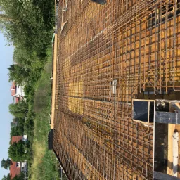 Zbrojenie fundamentu domu jednorodzinnego, widoczne drewniane szalunki i siatka z prętów stalowych, w tle zieleń i fragment zabudowy.