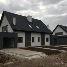 Dwa identyczne, dwukondygnacyjne domy z garażami, w trakcie wykańczania terenu przed budynkami; widoczne skrzynki elektryczne i gruz budowlany.